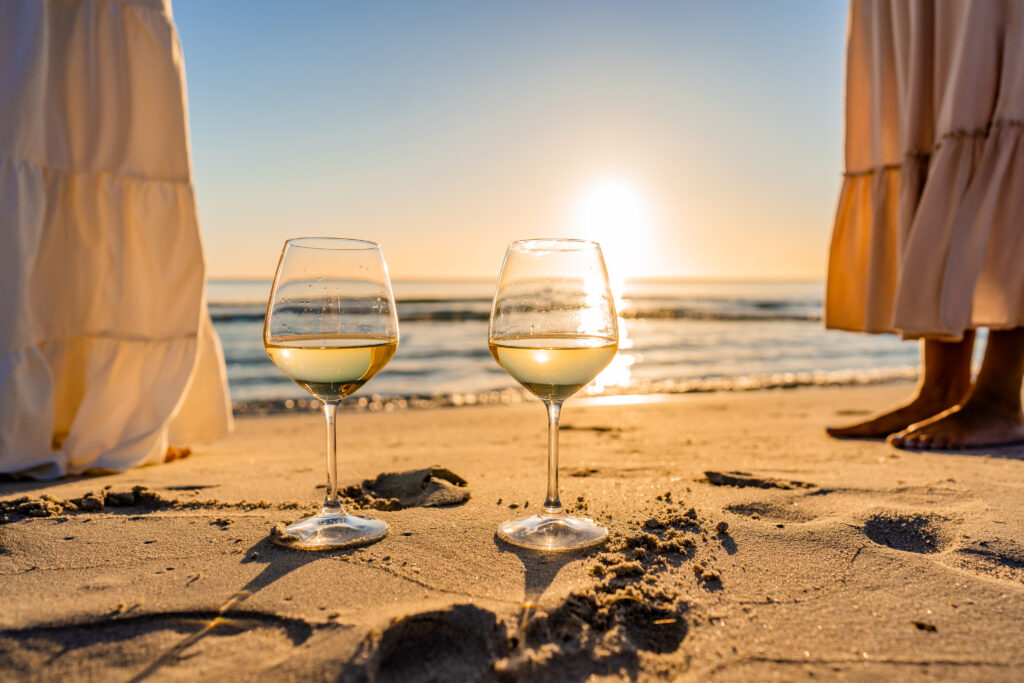 dos copas de vino blanco en la arena de la playa con una puesta de sol de fondo